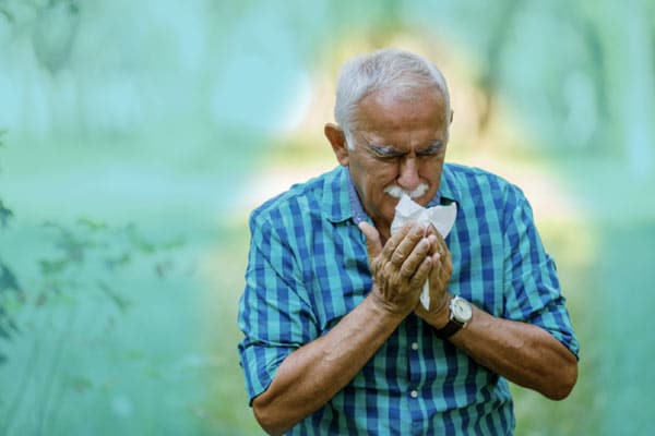 An older man sneezes as he stands outside.